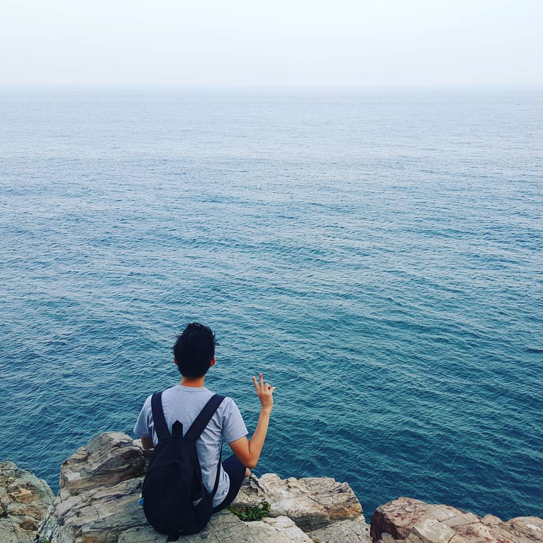 Endi 背后的一张照片,他坐在悬崖峭壁上,面向浩瀚的大海。他的右手做出 V 字手势。
